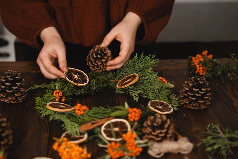 Hands arranging pine cones and dried oranges on a Christmas wreath.